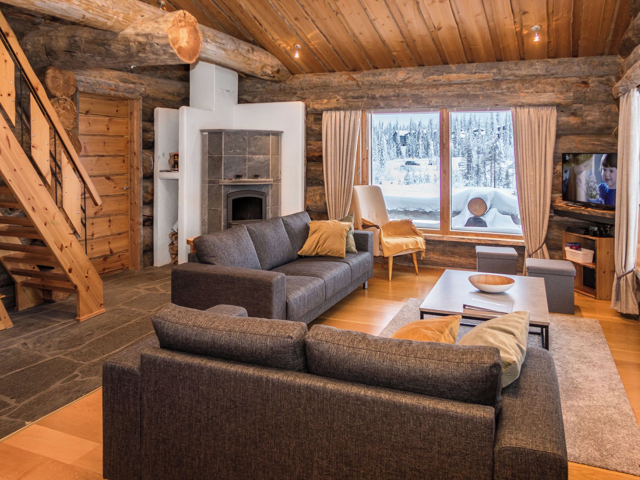 A log cabin interior with a fireplace, a sofa, a television, and a view of a snowy landscape.