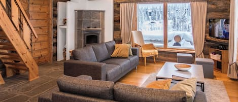 A log cabin interior with a fireplace, a sofa, a television, and a view of a snowy landscape.