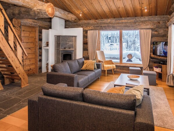 A log cabin interior with a fireplace, a sofa, a television, and a view of a snowy landscape.