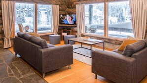 Ein gemütliches Blockhaus-Interieur mit zwei grauen Sofas, einem Couchtisch und einem Fernseher. Große Fenster bieten Blick auf eine verschneite Berglandschaft.