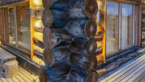 Eine Blockhütte mit einem zentralen Schornstein, großen Fenstern mit Jalousien und einer Holzterrasse.