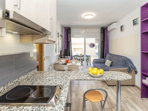 A compact kitchen with a built-in stove, a dining area with a table and chairs, a sofa, and a purple bookshelf.