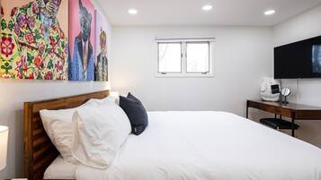 A hotel room with a large bed, a wooden headboard, a desk with a chair, a flat-screen TV, and a window.