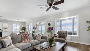 A spacious living room with a large window, a ceiling fan, and a wooden coffee table. - Kemah's Bayfront Getaway with Pool (Kemah)