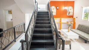 Una escalera con barandilla negra que lleva a un cuarto con una pared de acento naranja, un sofá blanco y una mesa con un florero.