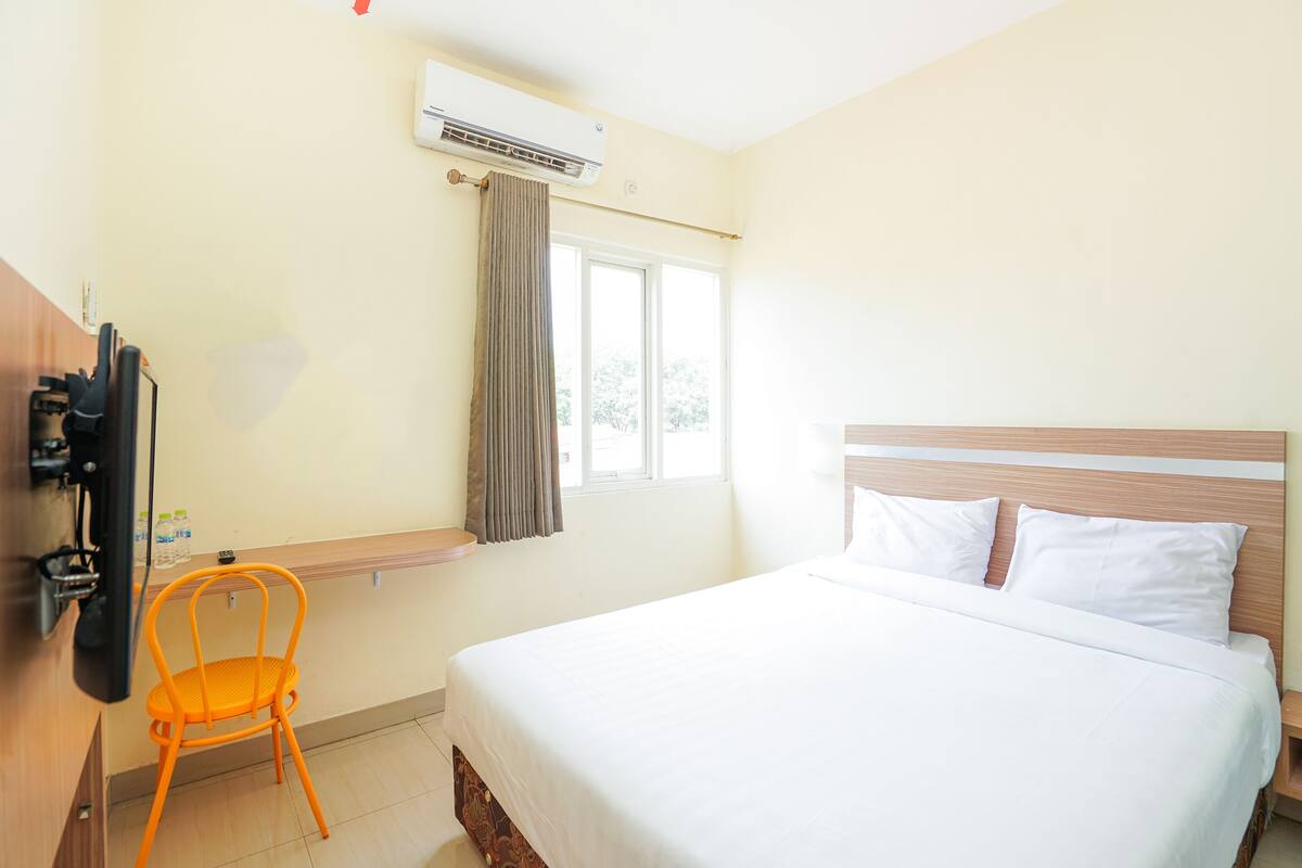 A hotel room with a bed, a chair, a TV, and an air conditioning unit.