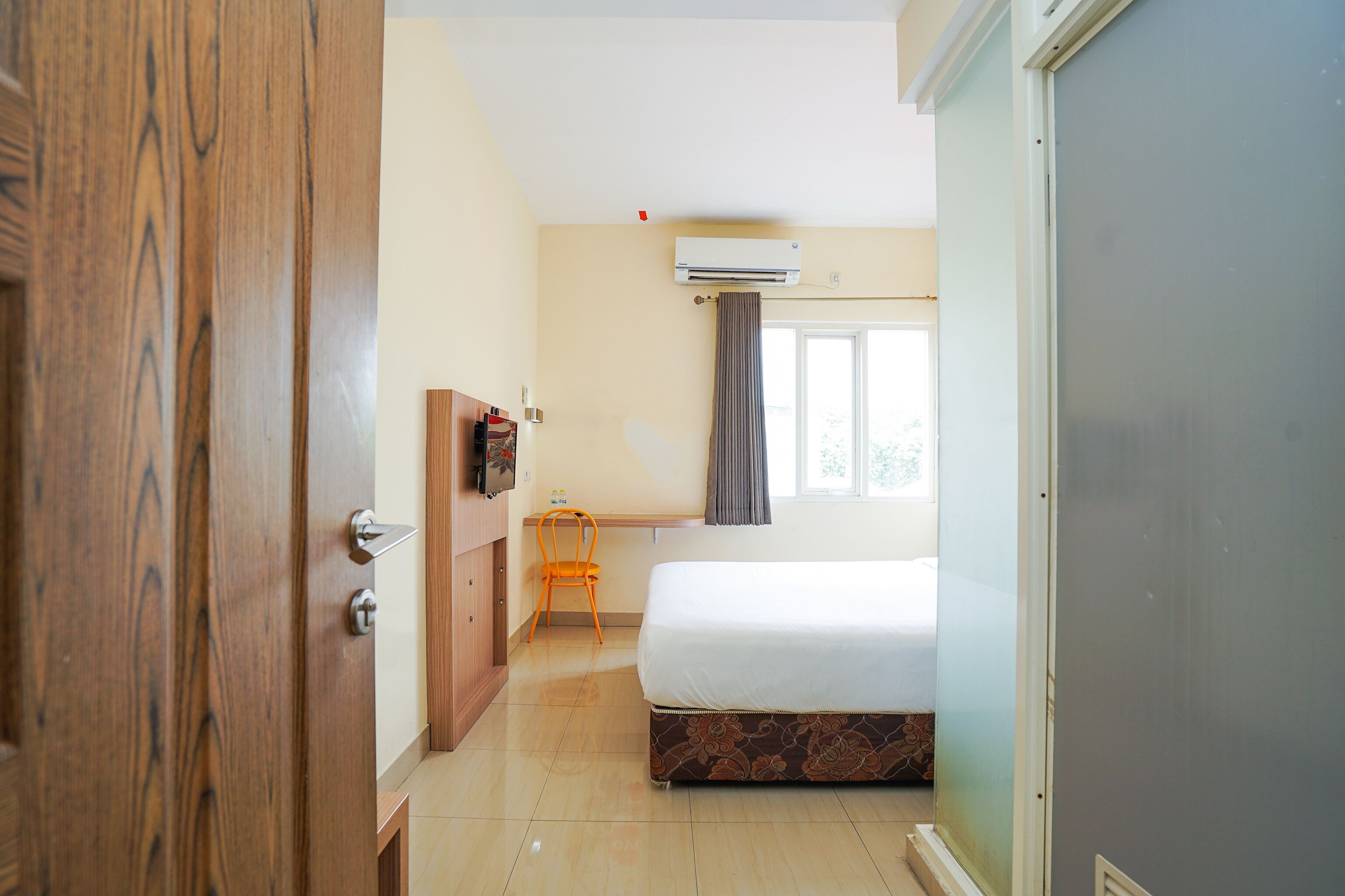 A hotel room with a wooden wardrobe, a desk with a chair, a bed with a patterned mattress, and an air conditioning unit.