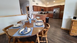 A dining area with a wooden table set for four, surrounded by wooden chairs. There is a kitchen area in the background with wooden cabinets and stainless steel appliances. - Beaverhead House Rental (Dillon)