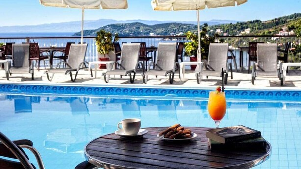 A poolside area with lounge chairs and umbrellas, a table with a drink and pastries, and a view of the sea and hills. - Poseidon Apartments (Skiathos)