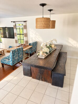 A modern living room with a wooden dining table, patterned chairs, a woven pendant light, and a TV mounted on the wall.