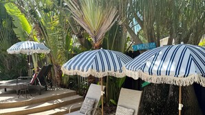 A sunlit outdoor seating area with striped umbrellas, lounge chairs, and a wooden table. - Just steps to the beach, sleeps 16, Golf cart, Dolphin Beach House huge pool! (Fort Lauderdale)