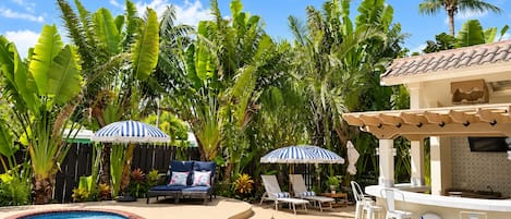 A swimming pool with a tiled edge, surrounded by lounge chairs and umbrellas, with palm trees in the background.