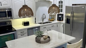 A modern kitchen with white cabinetry, a stainless steel refrigerator, a microwave, a toaster, and a sink. There is a marble island with two chairs and a decorative centerpiece. - Just steps to the beach, sleeps 16, Golf cart, Dolphin Beach House huge pool! (Fort Lauderdale)