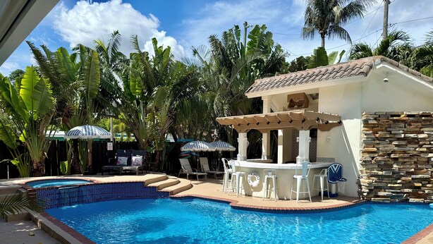 A swimming pool with lounge chairs and umbrellas, surrounded by tropical plants. - Just steps to the beach, sleeps 16, Golf cart, Dolphin Beach House huge pool! (Fort Lauderdale)