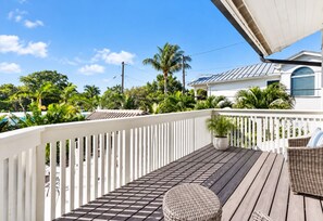 A wooden deck with a white railing, a potted plant, wicker furniture, and a house with a blue roof in the background.