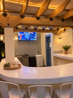 A bar area with a television, seating, and a kitchen in the background.