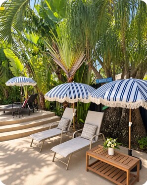 A tropical outdoor seating area with lounge chairs and striped umbrellas, surrounded by lush greenery. - Just steps to the beach, sleeps 16, Golf cart, Dolphin Beach House huge pool! (Fort Lauderdale)