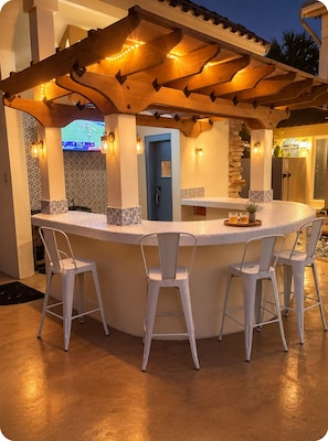 An outdoor bar area with a curved counter, three white bar stools, and a stone pillar.