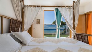 Un lit recouvert d’une couvre-lit à motifs, une chaise en osier et une vue sur l’océan par une fenêtre ornée de rideaux.