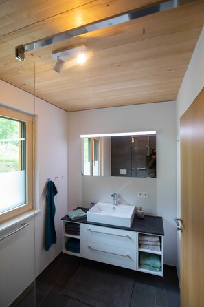 Un baño moderno con techo de madera, un espejo grande, un lavabo blanco y una ventana.