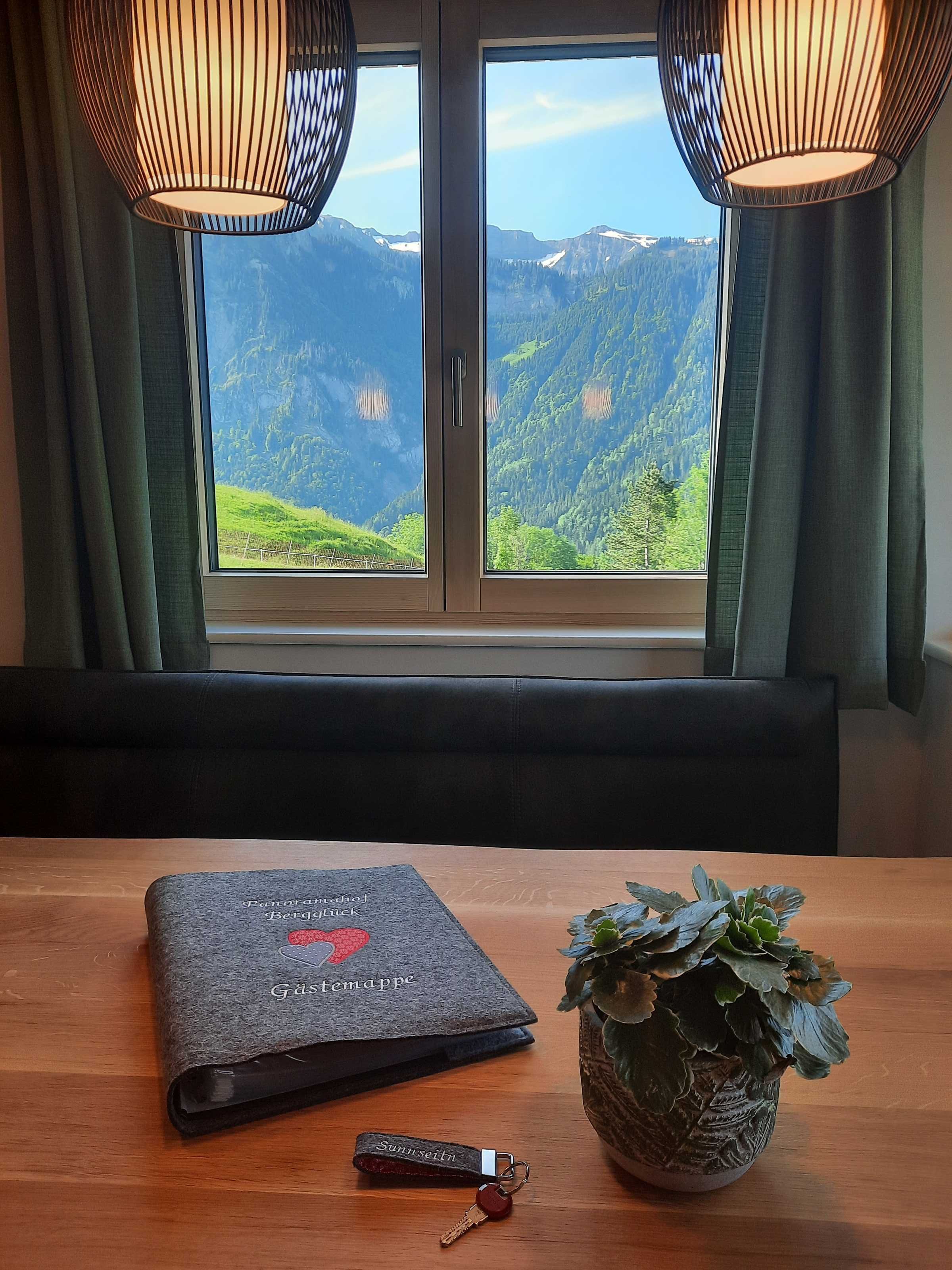 Ein Zimmer mit einem großen Fenster, einem Tisch mit einem Buch und einem Schlüsselanhänger, sowie einer Topfpflanze.