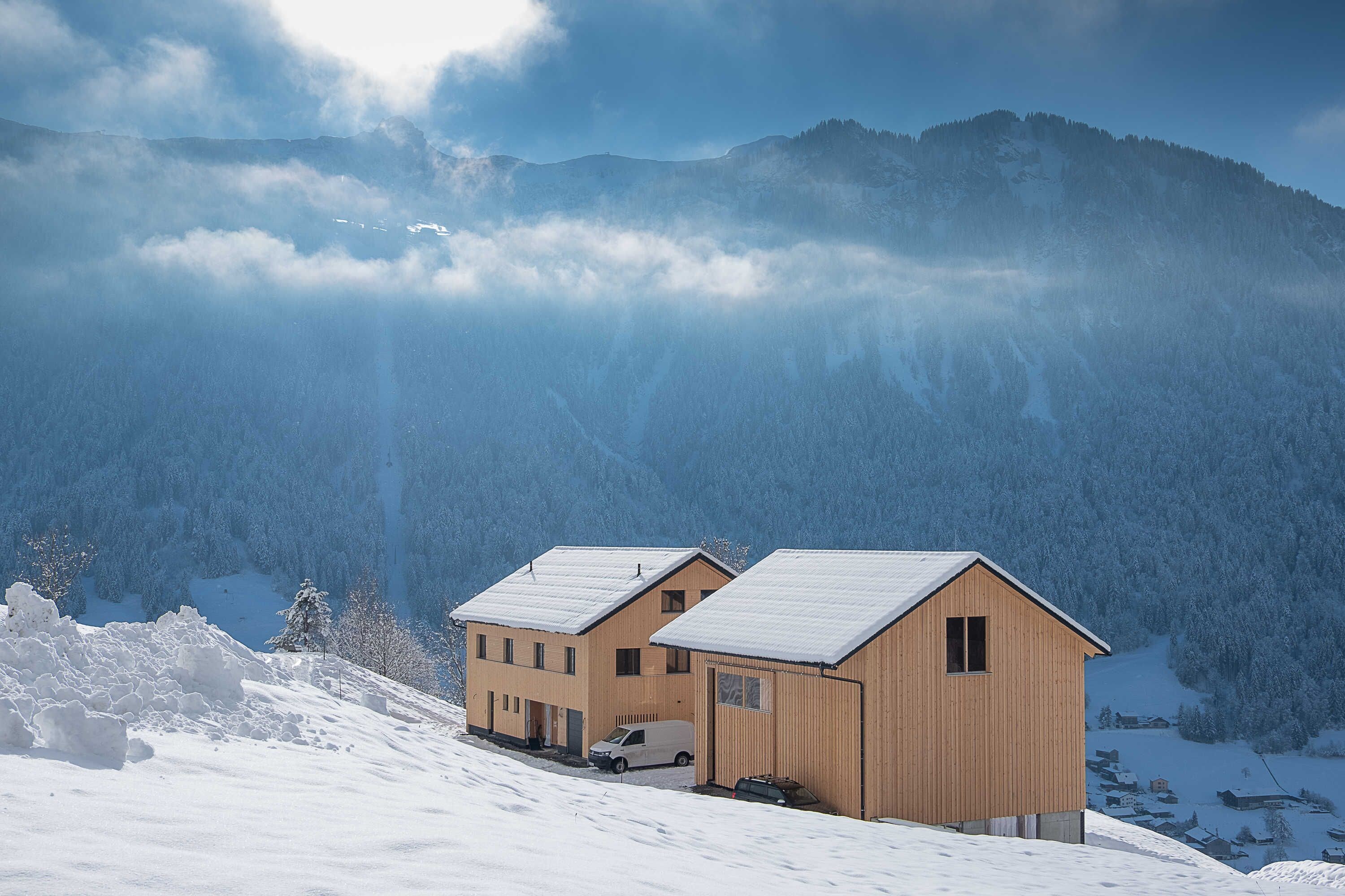 A two-story wooden chalet with a snow-covered roof, situated on a snowy mountain slope with forested background.
