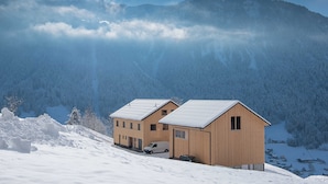 A two-story wooden chalet with a snow-covered roof, situated on a snowy mountain slope with forested background. - Vacation apartment Sunnseitn - Panoramahof Bergglück (Mellau)