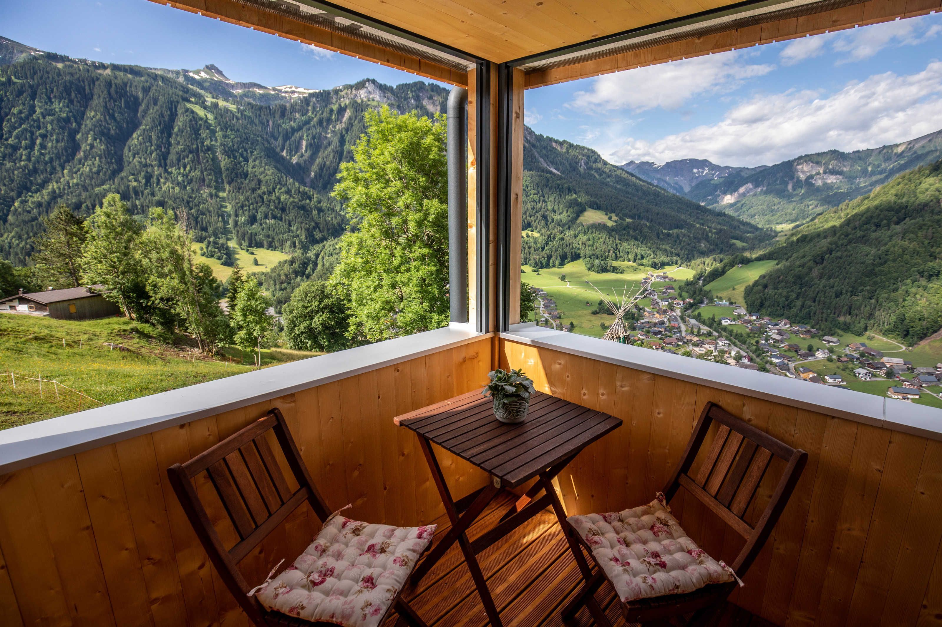 Ein Balkon mit Holzmöbeln, einem kleinen Tisch und Blick auf eine gebirgige Landschaft.