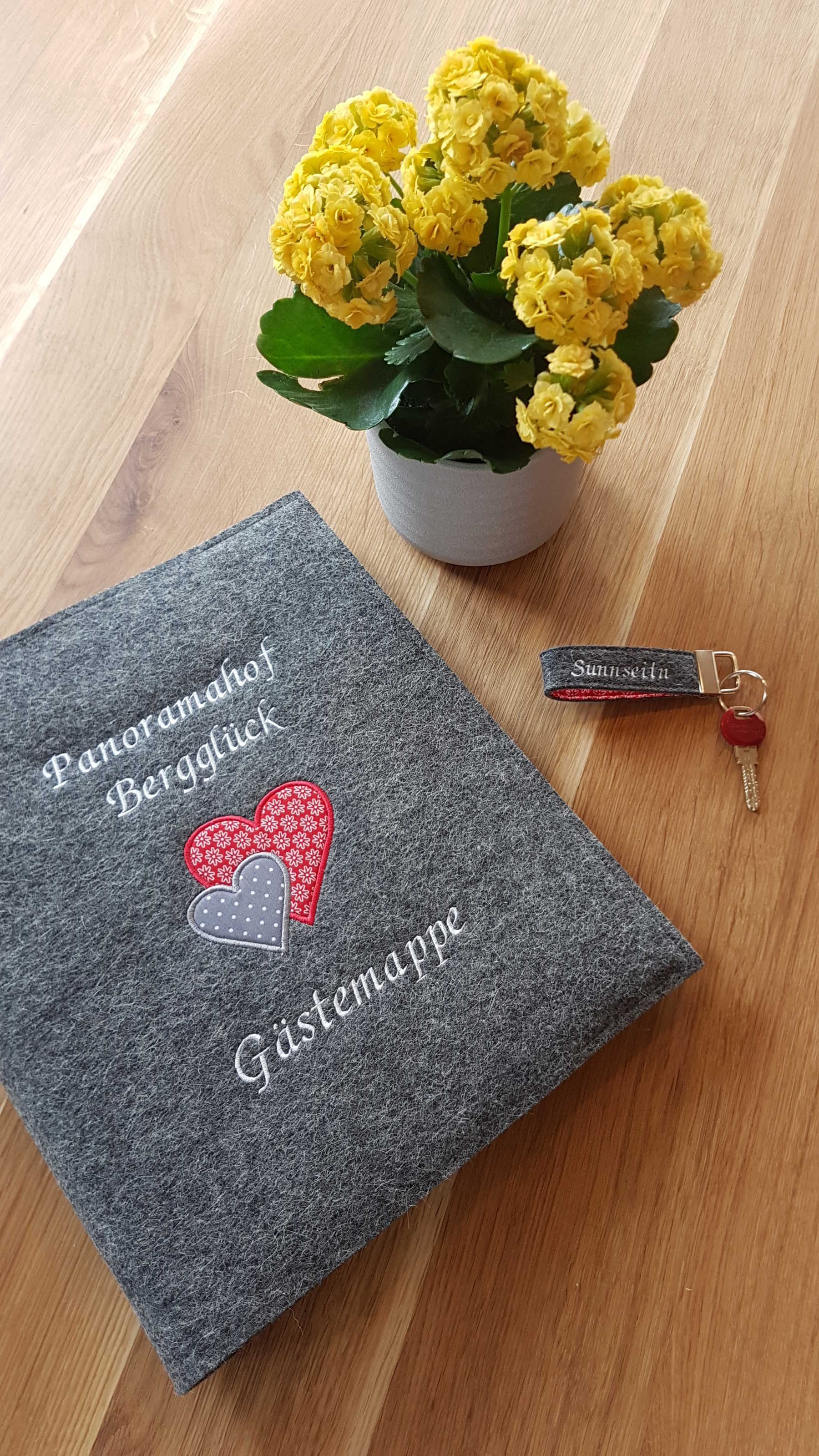 Un libro de fieltro gris con texto bordado y un corazón, una flor amarilla en maceta y un llavero sobre una superficie de madera.