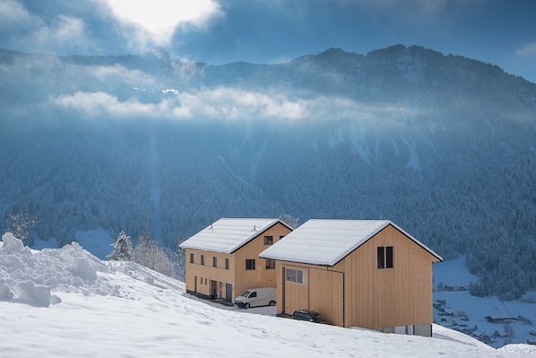 A two-story wooden chalet with a snow-covered roof, situated on a snowy mountain slope with forested background.