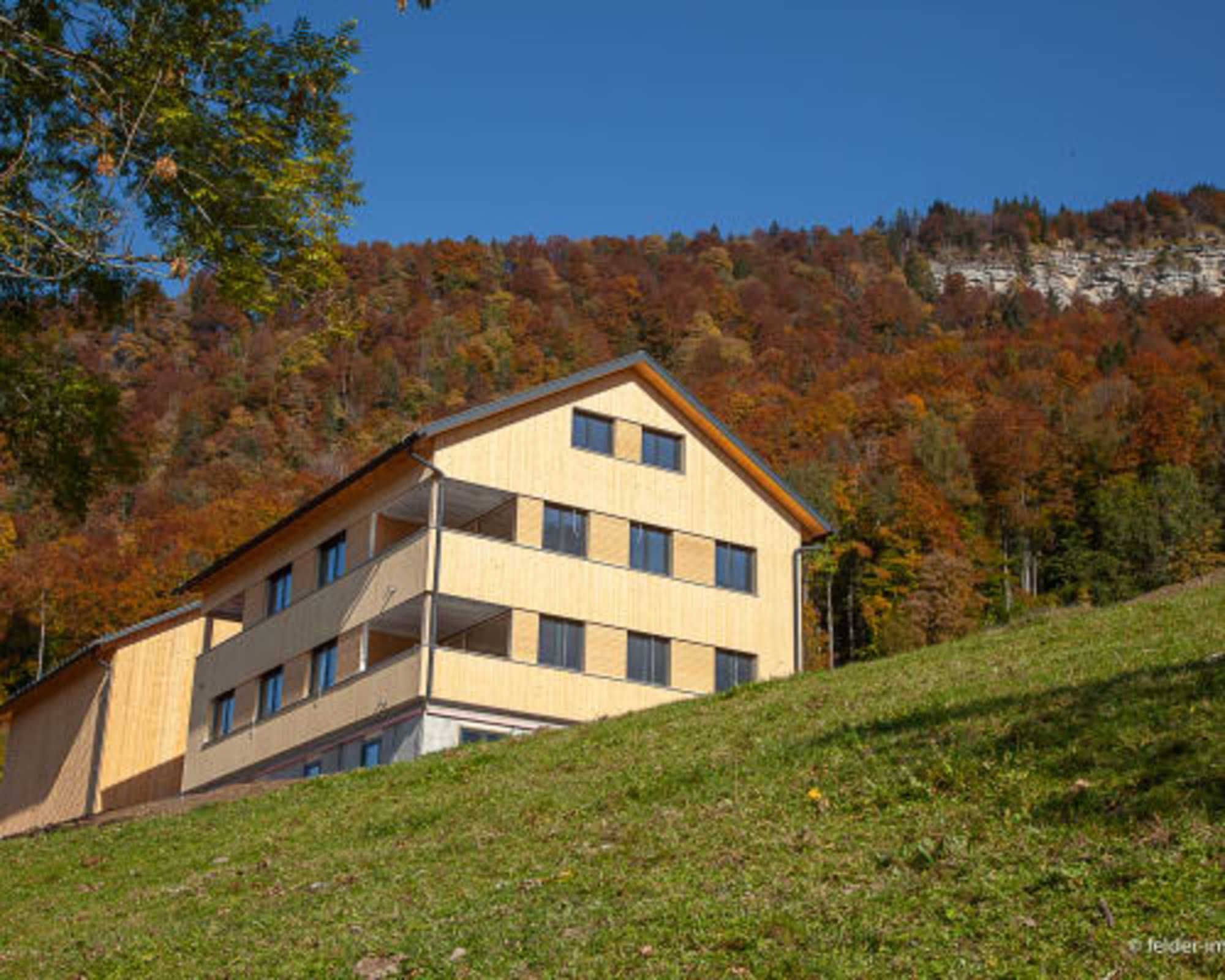 A multi-story building with a flat roof, situated on a grassy slope with a forested hillside in the background.