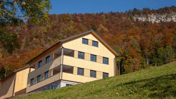 A multi-story building with a flat roof, situated on a grassy slope with a forested hillside in the background.