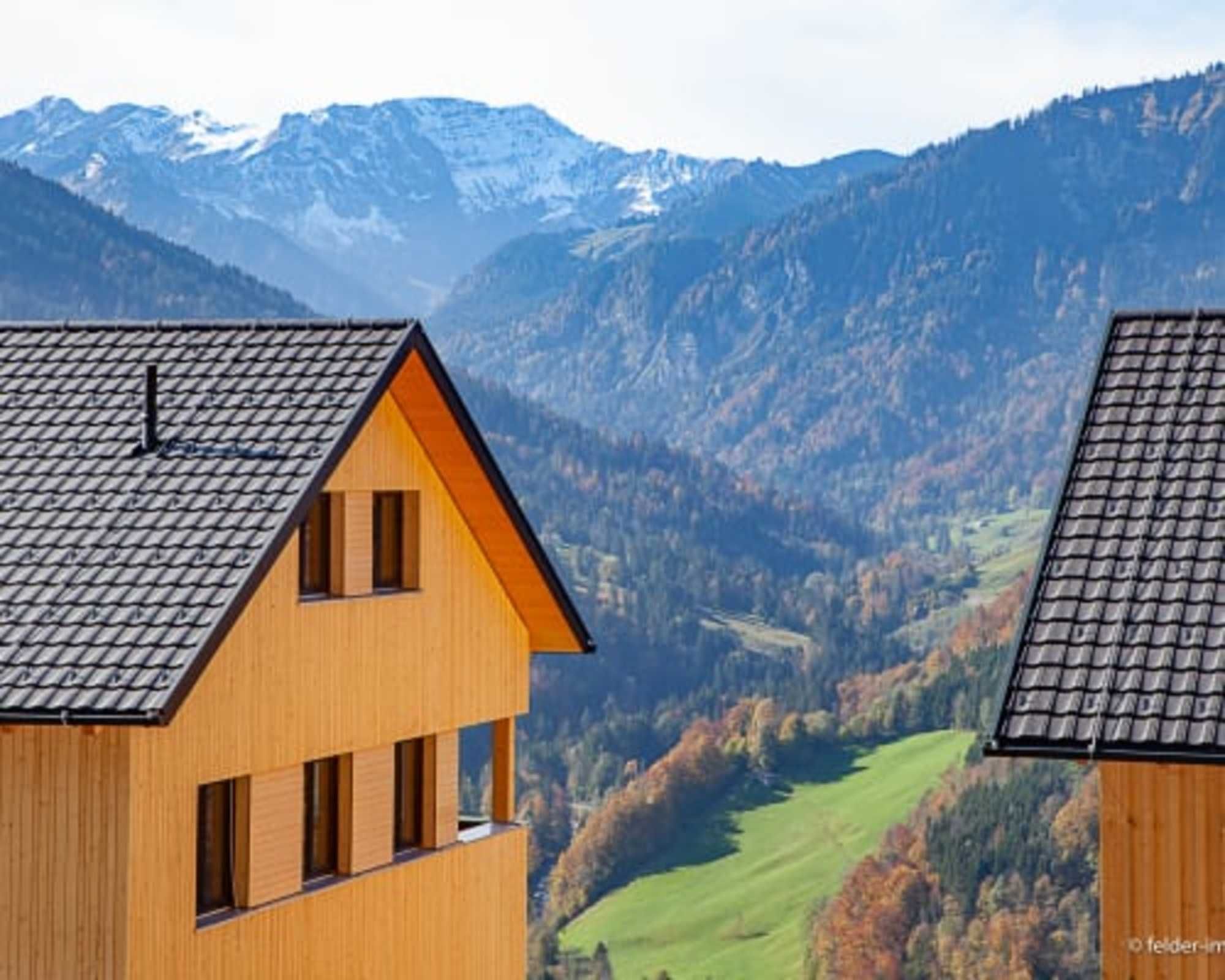 A wooden house with a sloped roof, a balcony, and a view of a valley with mountains in the background.