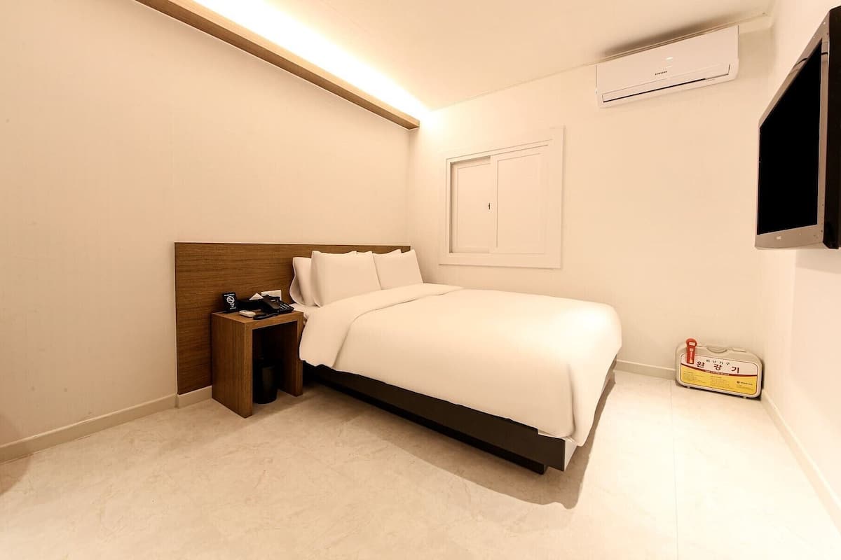 Ein modernes Hotelzimmer mit einem ordentlich bezogenen Bett, einem Nachttisch mit Telefon und Taschentüchern, einem an der Wand montierten Fernseher und einem kleinen zusätzlichen Nachttisch mit einer Flasche und einem Schild.