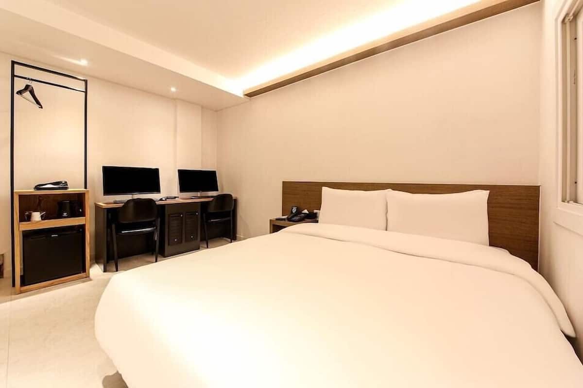 Ein Hotelzimmer mit einem großen Bett, einem Schreibtisch mit zwei Monitoren, einem Stuhl und einem Kleiderschrank.