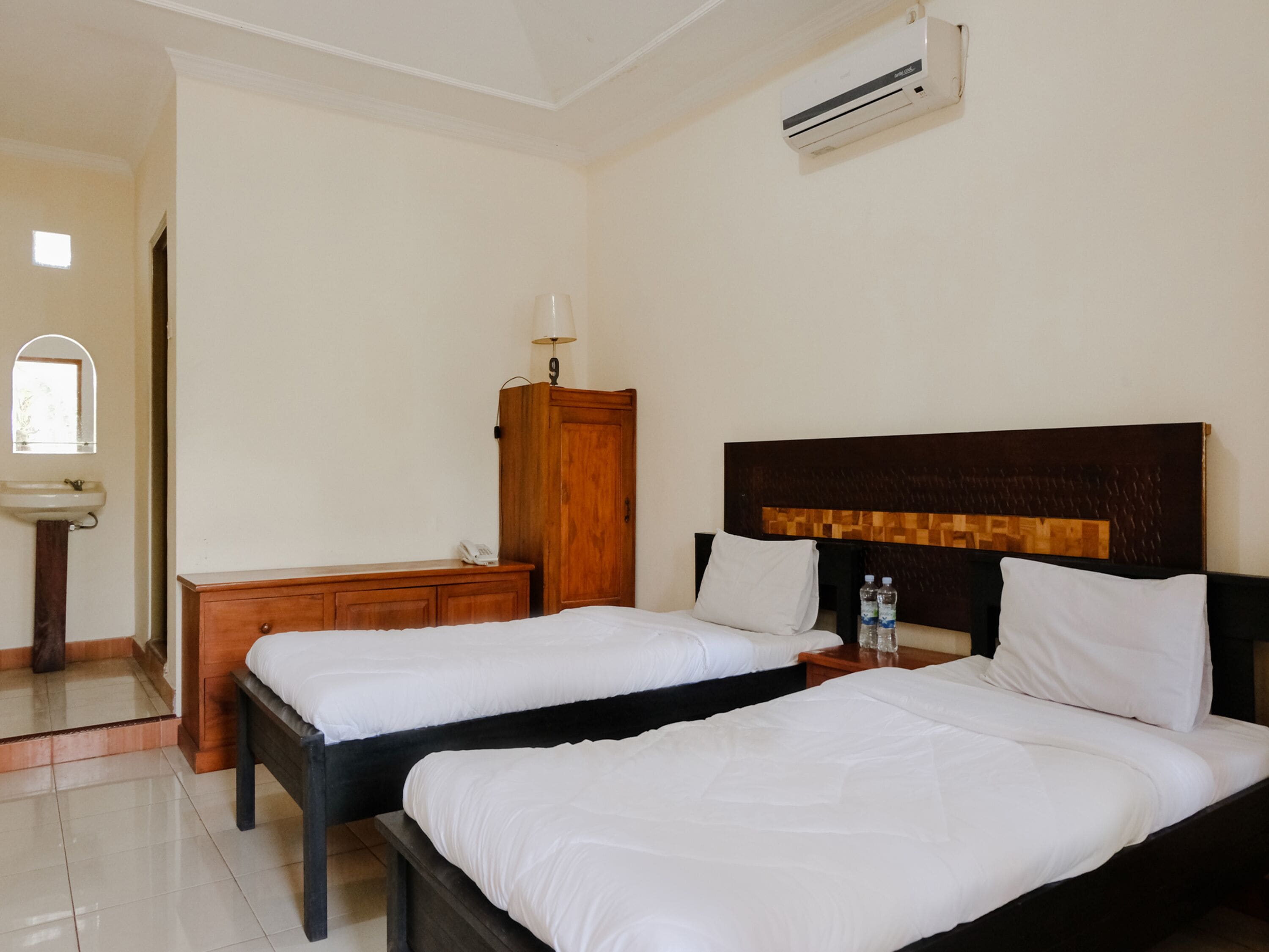 Une chambre d’hôtel avec deux lits, une armoire en bois, une table de chevet avec une lampe, un petit lavabo et un appareil de climatisation.