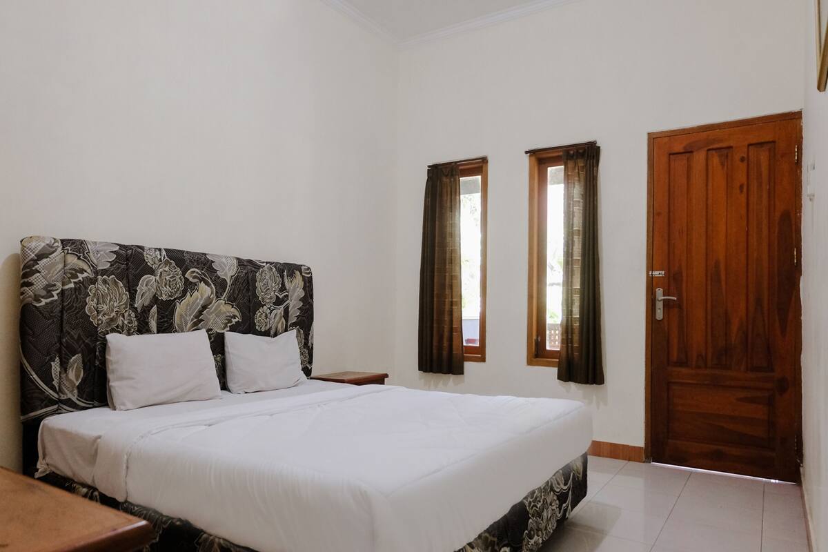 A bedroom with a bed, two windows, a wooden door, and a nightstand.