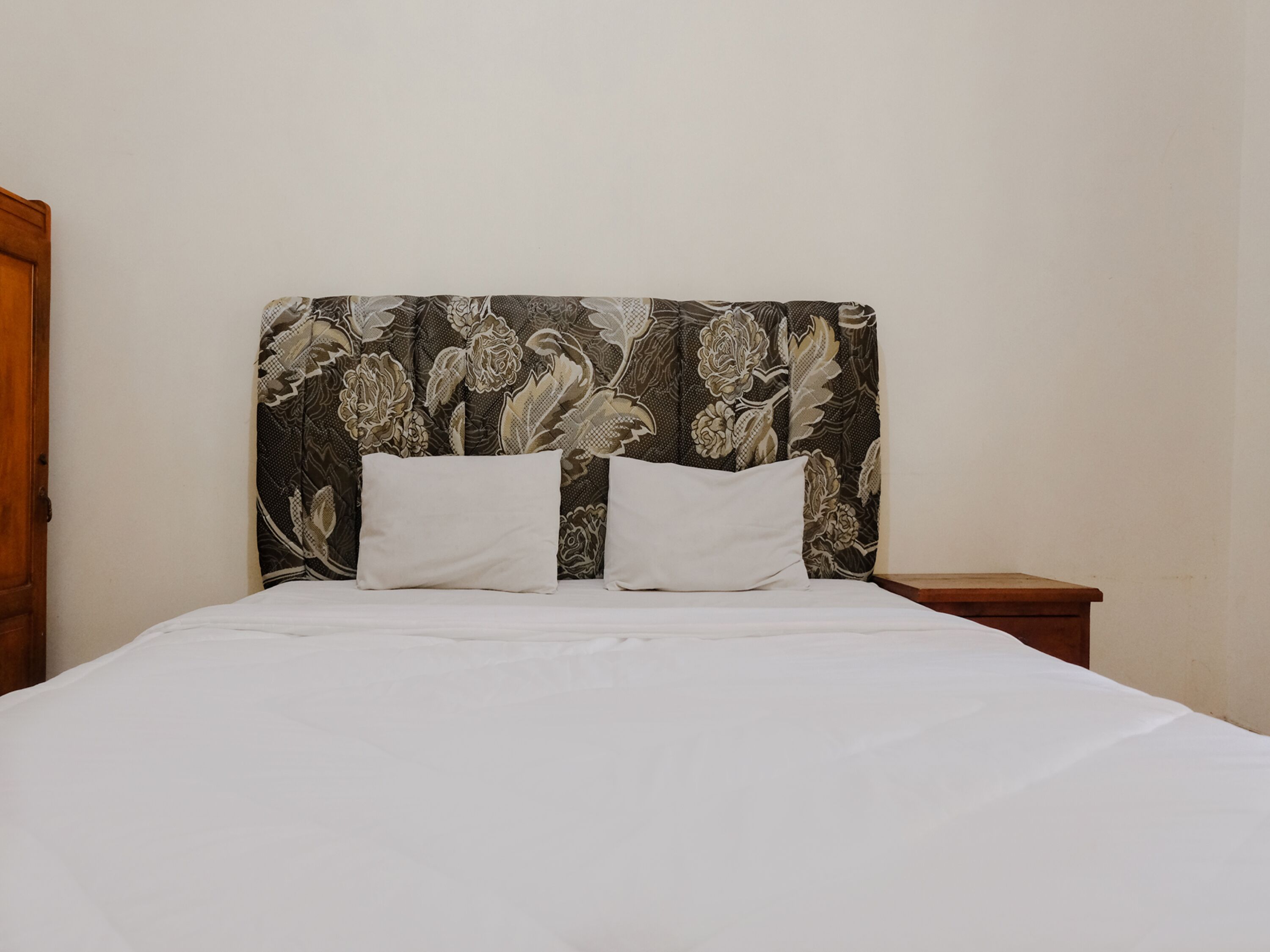 A bed with white bedding and two pillows, a wooden nightstand, and a headboard with a floral pattern.