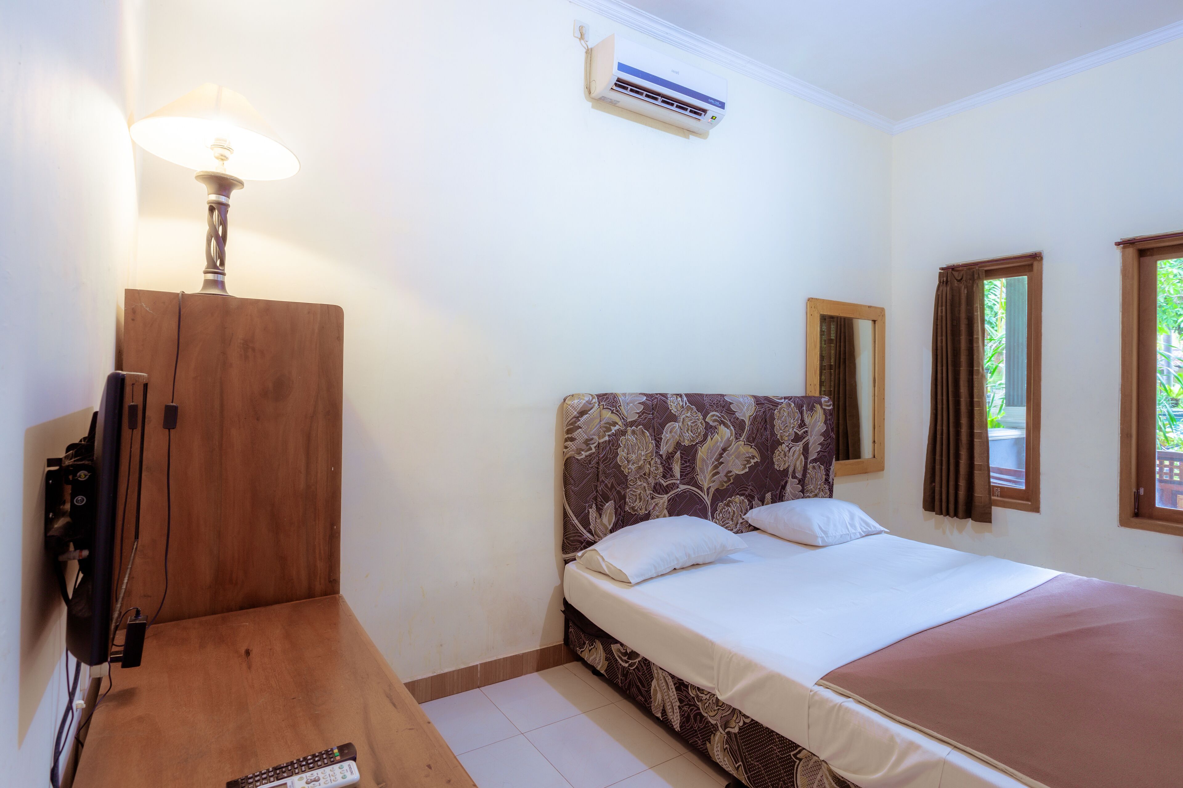 A bedroom with a bed, a wooden nightstand, a lamp, an air conditioner, and a window with curtains.