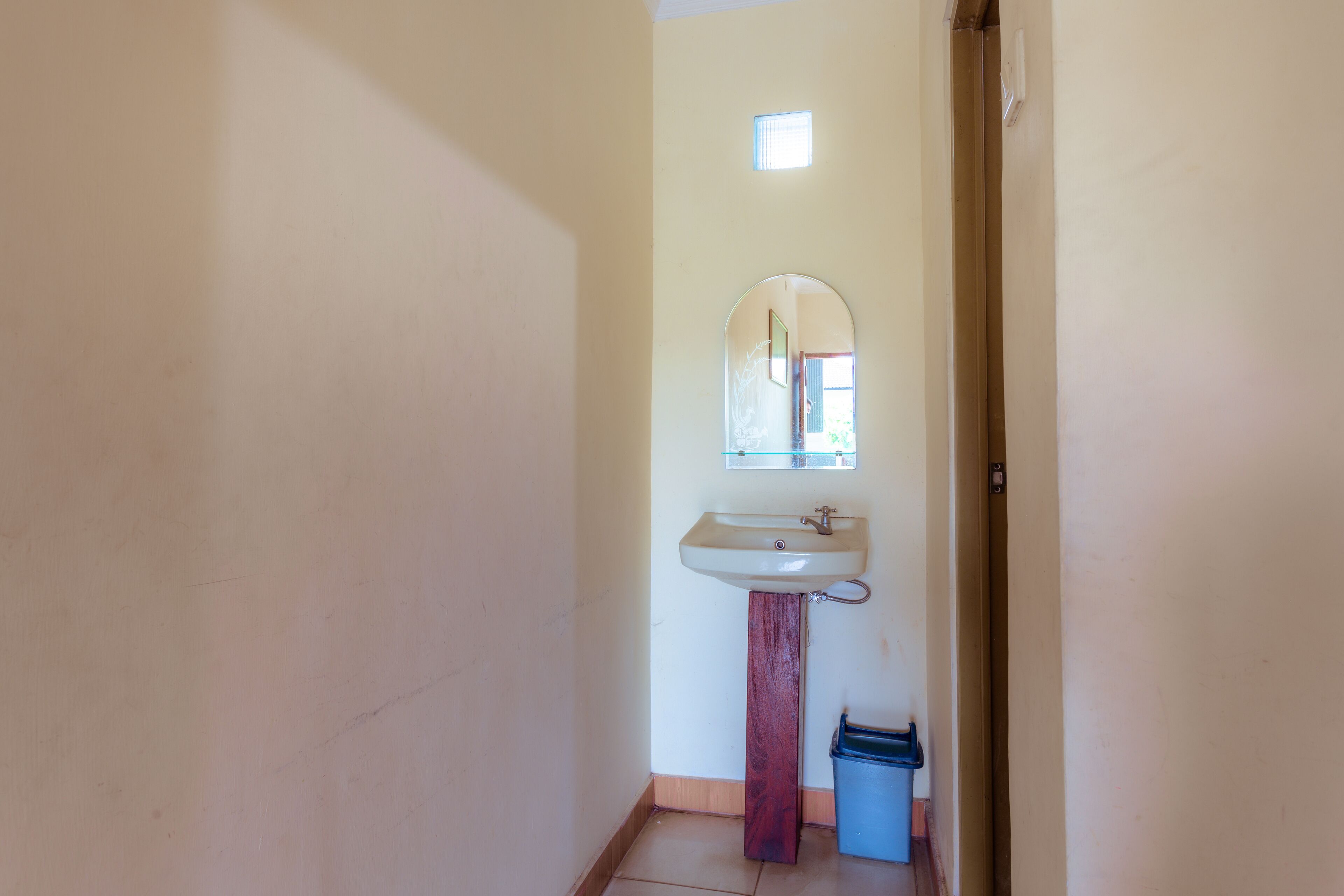 A narrow hallway with a white sink and mirror on the right, a small window, and a blue trash bin.