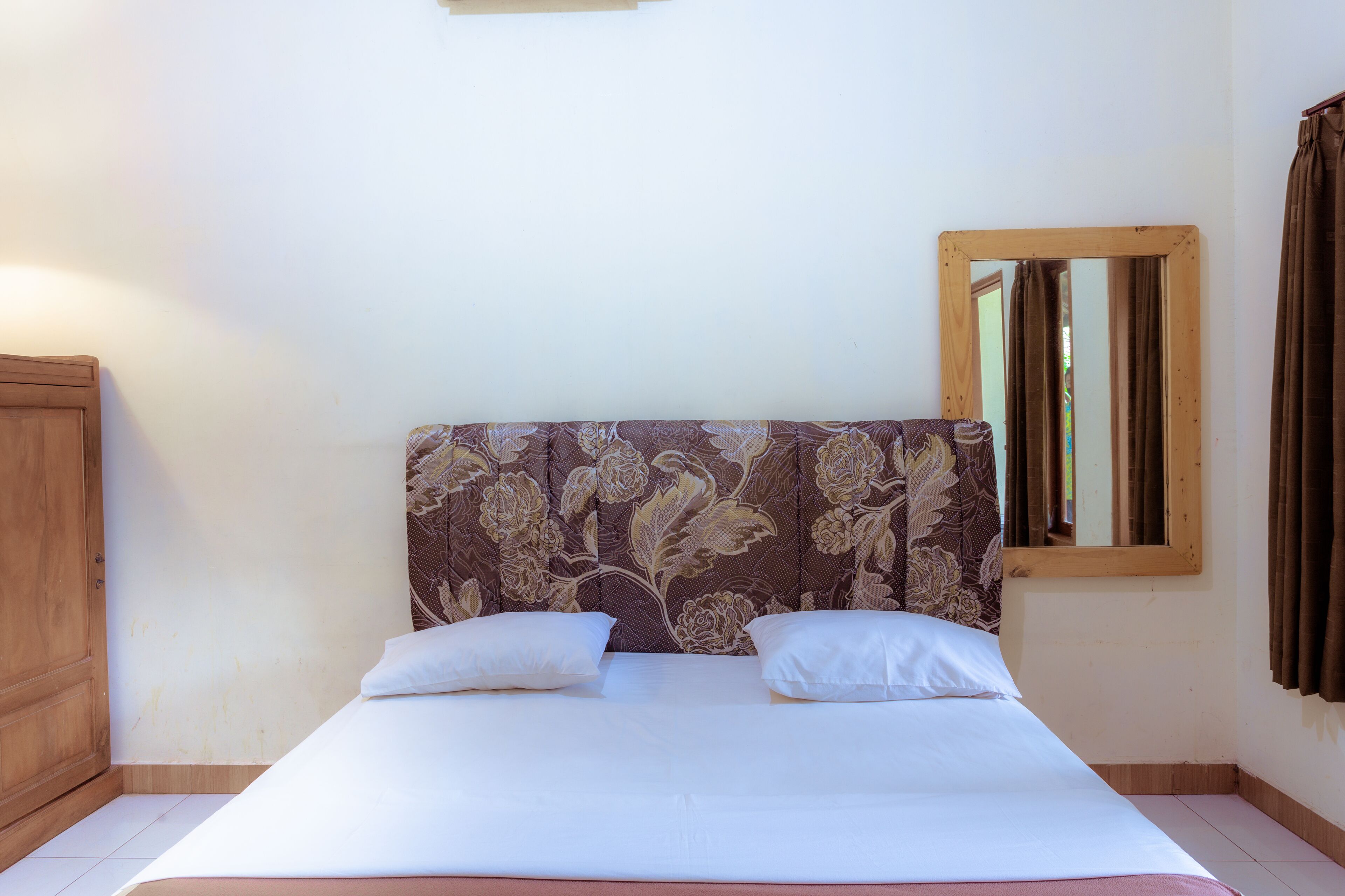 A bedroom with a bed, two pillows, a wooden headboard, a mirror, and a curtain.