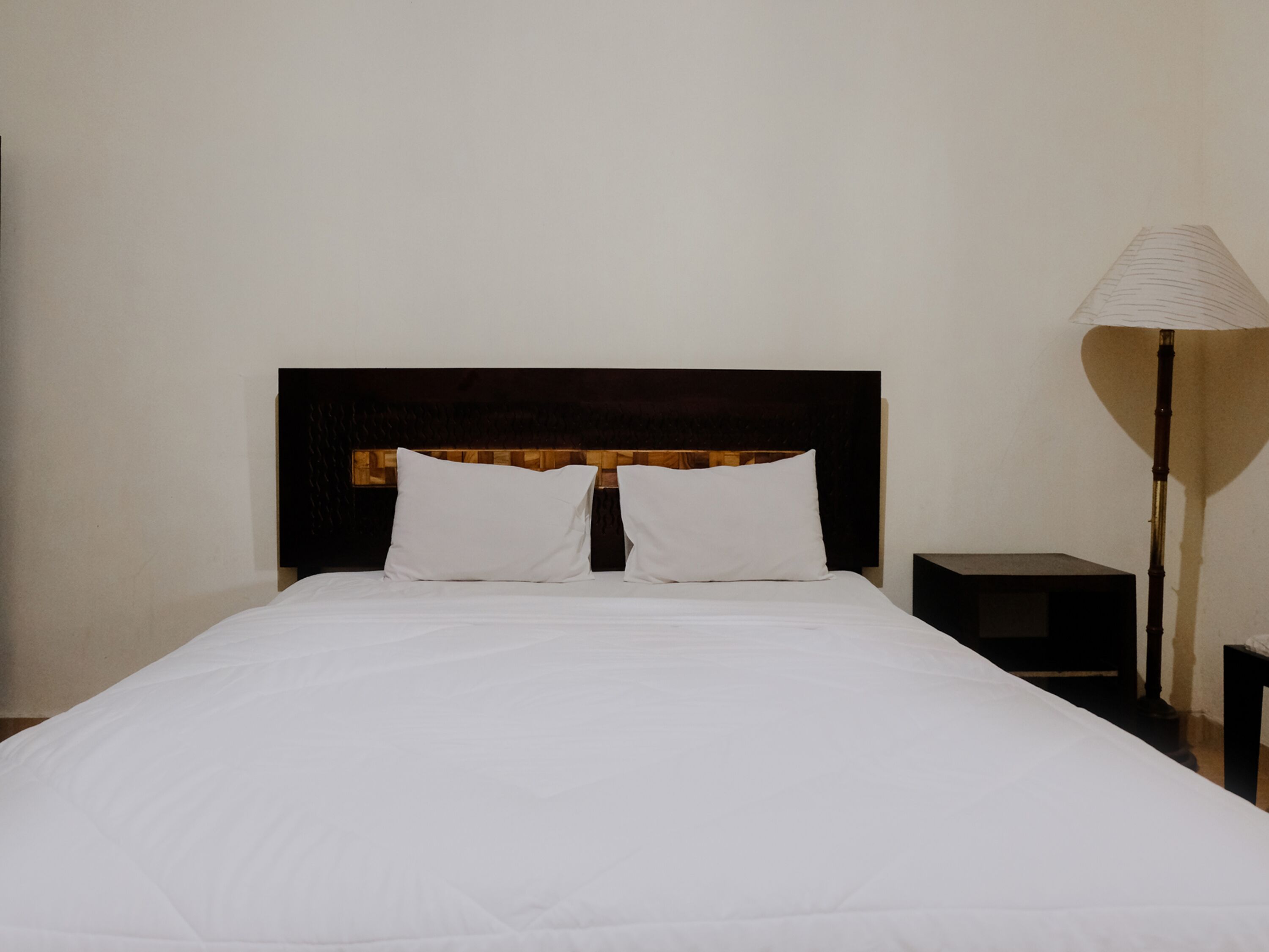 A neatly made bed with white linens, a dark wooden headboard, and a bedside lamp.