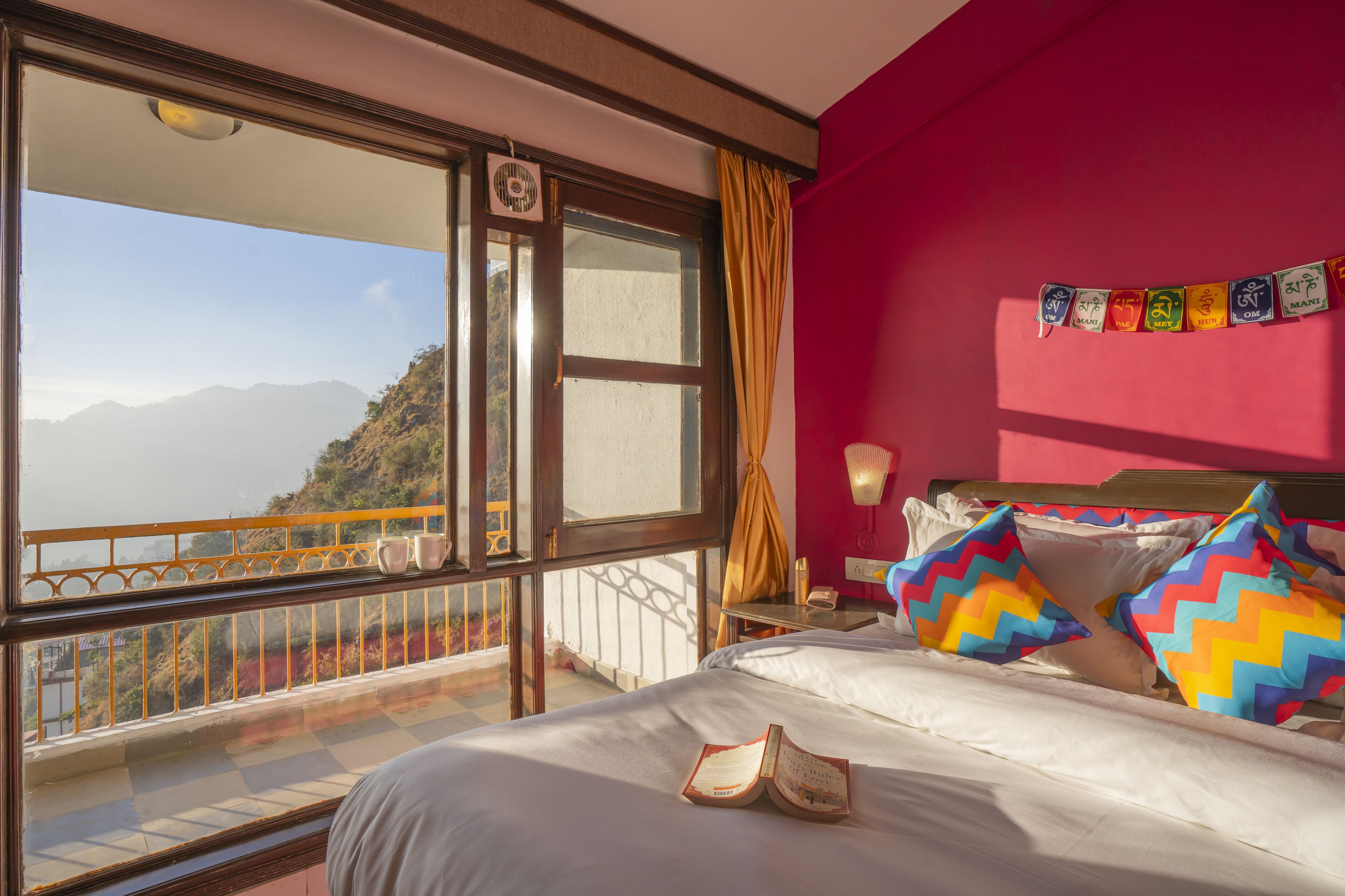 A hotel room with a large window, a bed with colorful pillows, a bedside table with a lamp, and a wall with framed pictures.