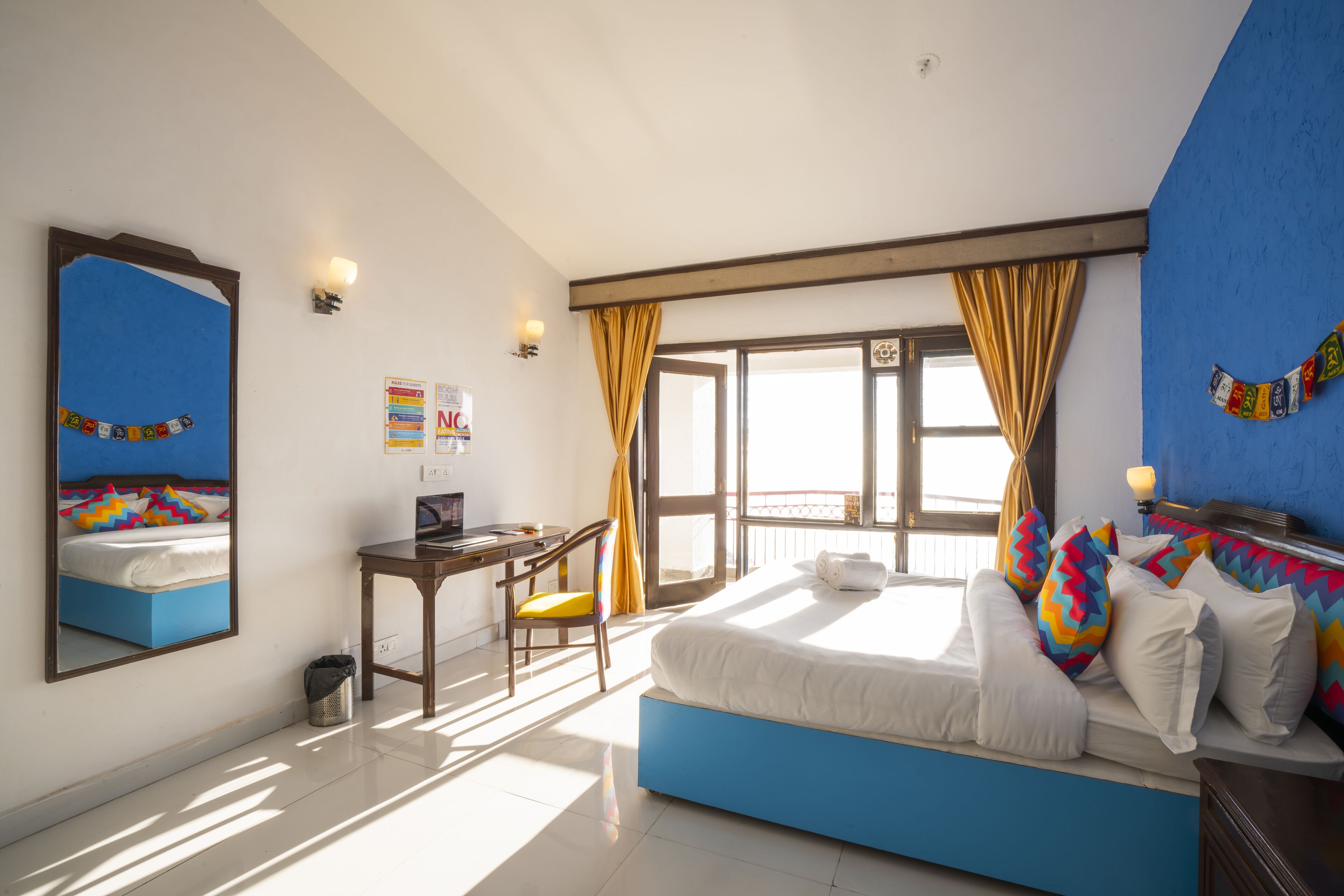 A bedroom with a bed, a desk, a chair, a window with curtains, and a mirror on the wall.