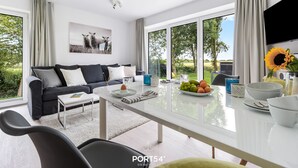 Ett modernt vardagsrum med en soffa, ett soffbord och ett matbord. Det finns stora fönster med gardiner, en platt-TV och en vas med solrosor.