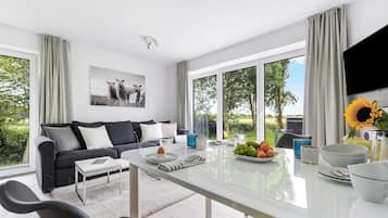 Una sala de estar moderna con sofá, mesa de centro y comedor. Hay ventanas amplias con cortinas, un televisor de pantalla plana y un jarrón con girasoles.
