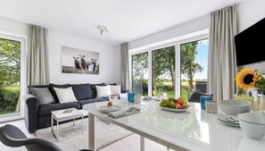 A modern living room with a sofa, coffee table, and dining table. There are large windows with curtains, a flat-screen TV, and a sunflower vase. - Grünwald – for 4 people, 30 minutes from Sylt, idyllic and absolutely peaceful. (Emmelsbüll-Horsbüll)