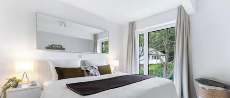 Ein modernes Schlafzimmer mit einem großen Bett, einem Nachttisch mit Lampe, einem Spiegel und Blick ins Grüne.
