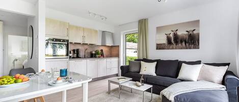 A modern living room with a dark grey sofa, a coffee table, and a dining area with a white table and fruit bowl.