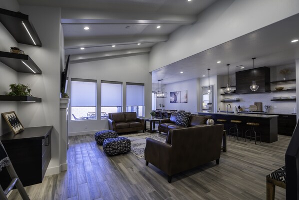 A modern living room with a sofa, ottoman, and coffee table, a dining area with chairs, and a kitchen with bar stools.